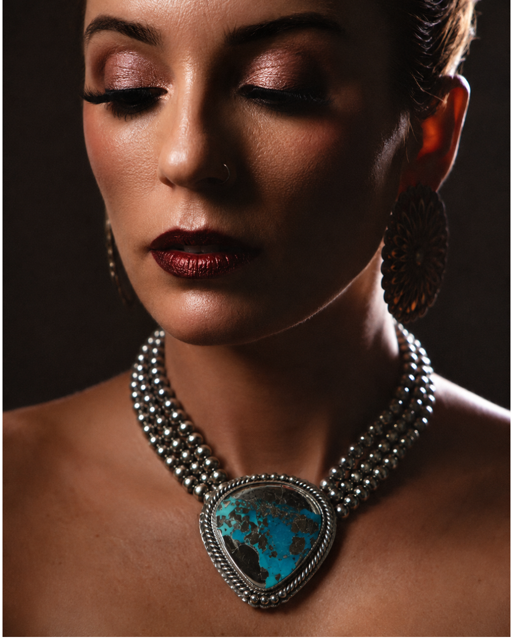 A close-up portrait of a woman wearing a silver-toned multi-strand necklace with a large turquoise and black stone pendant, against a dark brown background.
